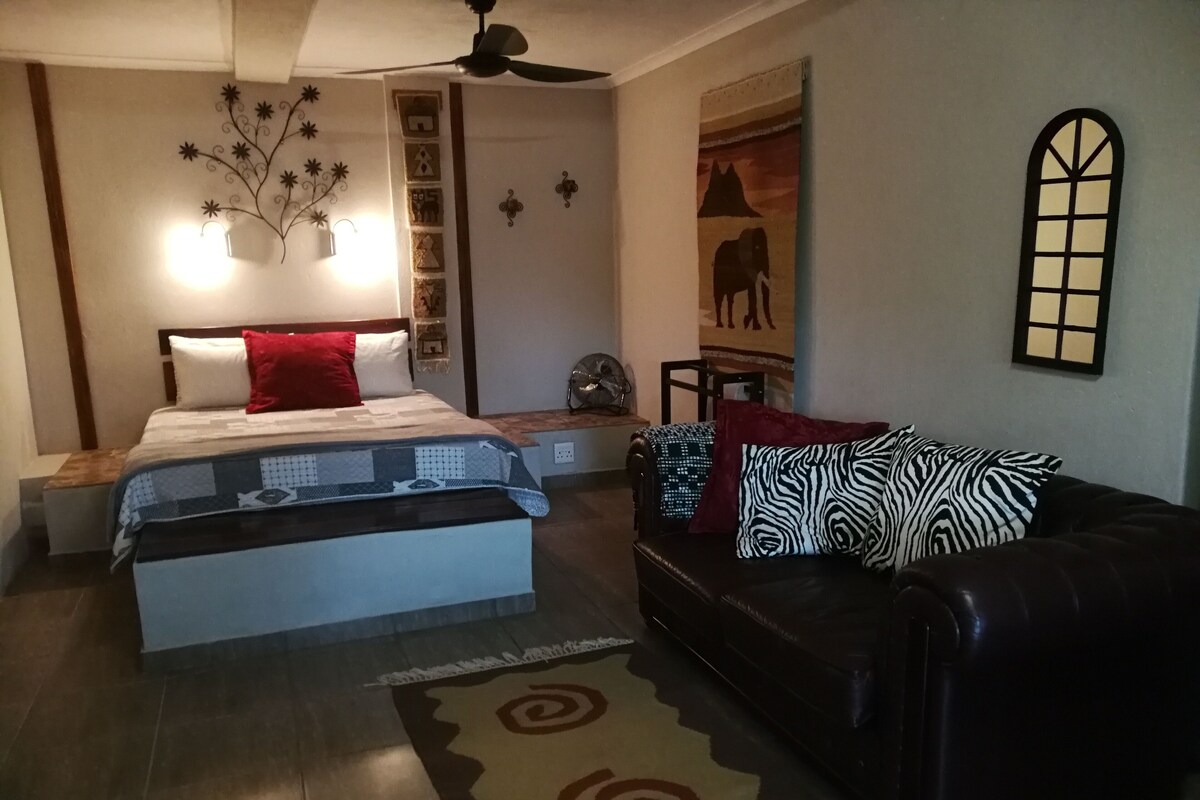 A bedroom with a bed, a black leather sofa, a rug, a ceiling fan, and wall decorations.
