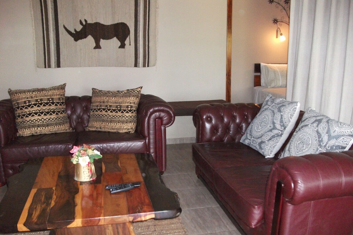 A living room with a dark brown leather sofa, two patterned throw pillows, a wooden coffee table with a remote control, and a wall-mounted artwork of a rhinoceros.