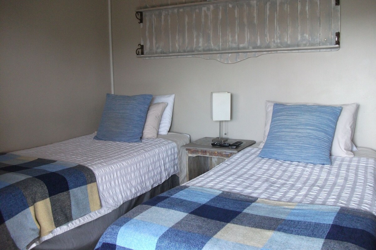 A hotel room with two beds, each with a checkered blanket, a nightstand with a lamp, and a shelf above the beds with potted plants.