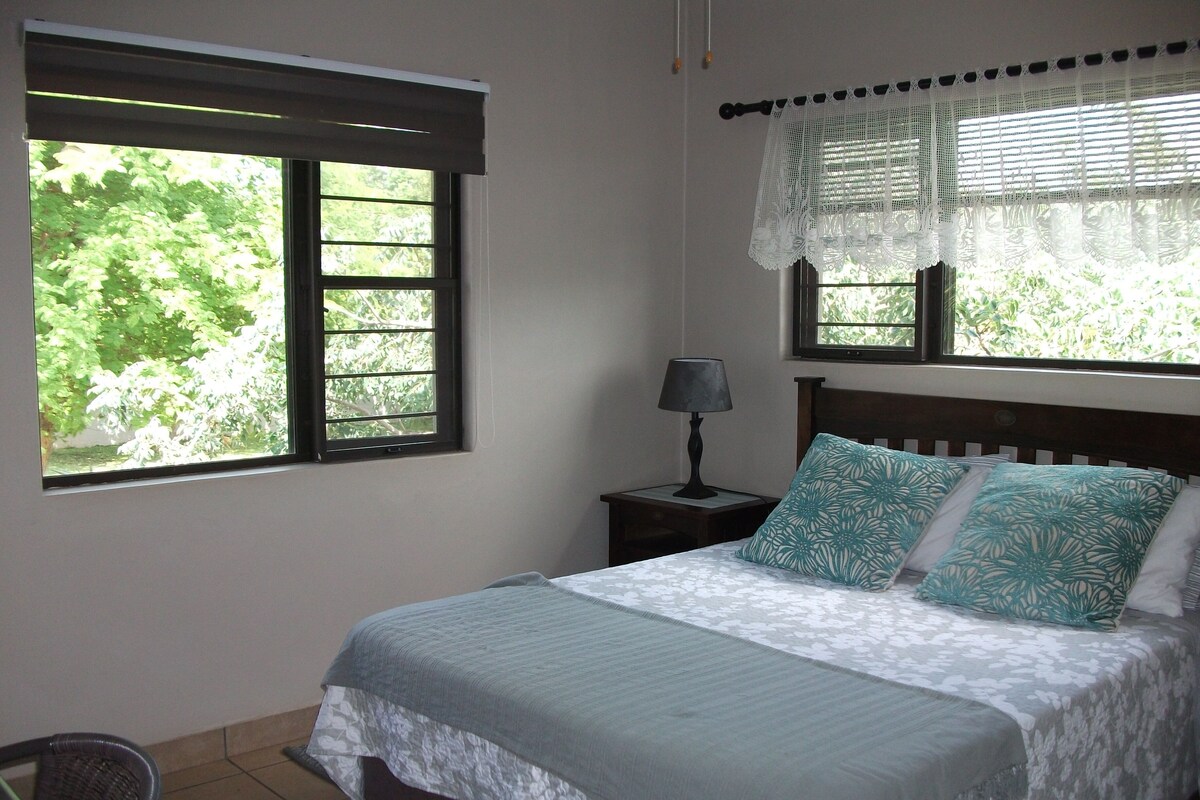 A bedroom with a bed, two pillows, a nightstand, a lamp, and two windows with blinds.