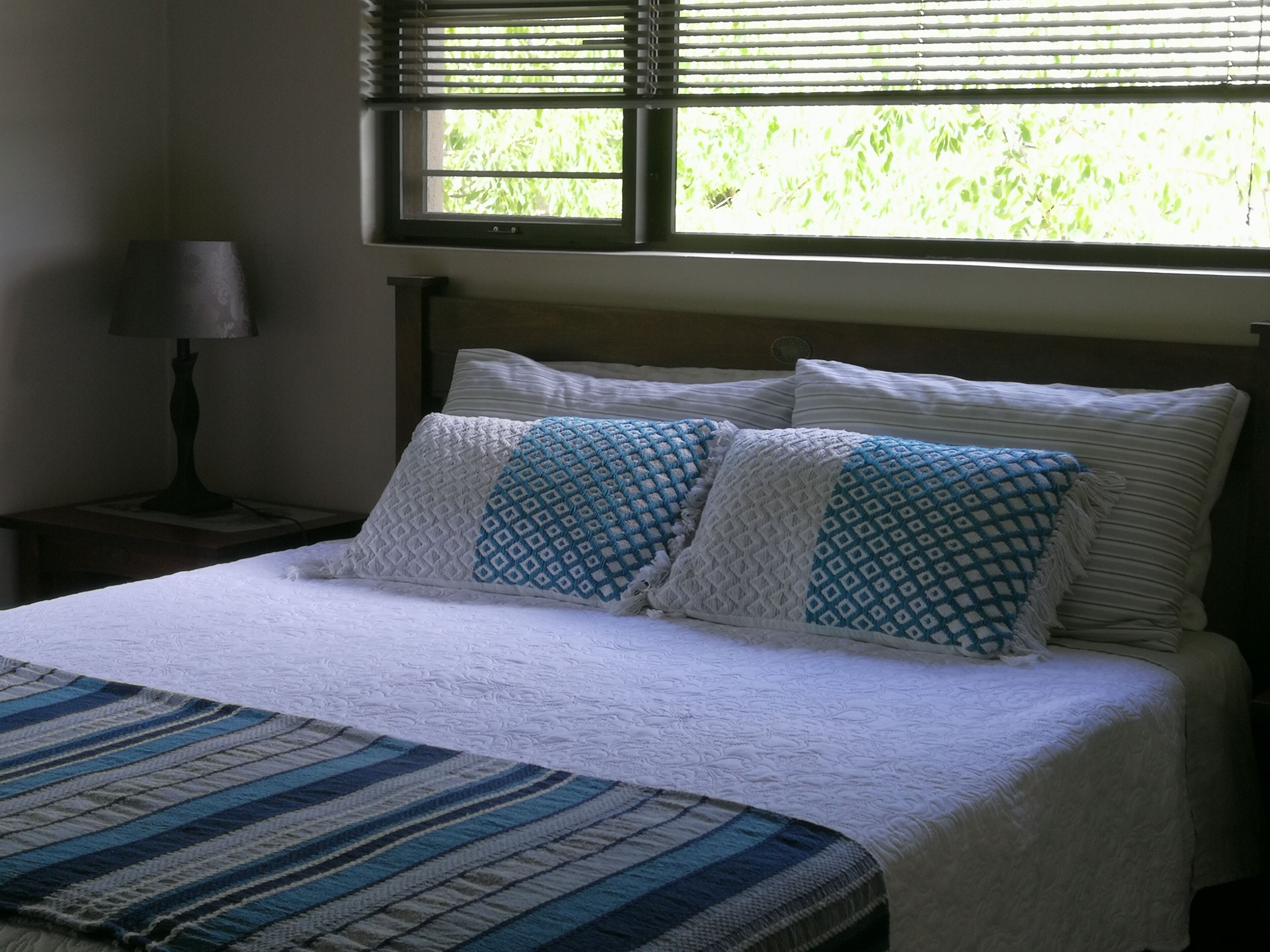 A bedroom with a bed, pillows, a nightstand, a lamp, and a window with blinds.