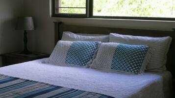 A bedroom with a bed, pillows, a nightstand, a lamp, and a window with blinds.