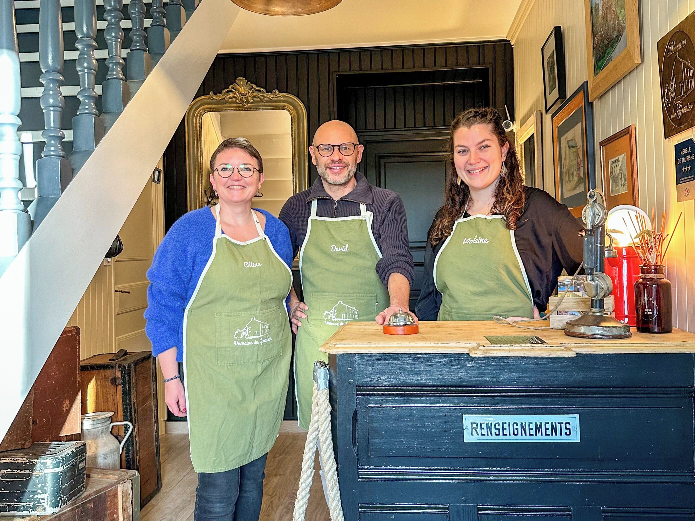 Drie personen staan in een hal met een trap aan de linkerkant, een houten balie met een bord waarop 'Renseignements' staat op de voorgrond, en een deur met een spiegel en ingelijste foto's aan de muur.