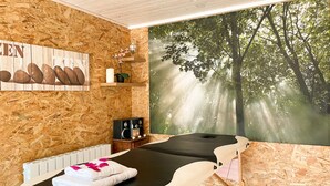 A massage room with a wooden wall, a massage table, a radiator, a shelf with decorative items, and a large window with a forest view. - Domaine du Grenier (Yffiniac)