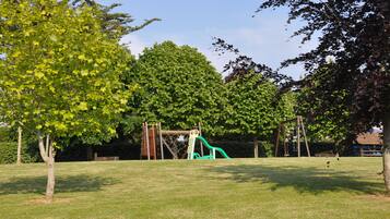 Children can play on the playground's green slide and climbing structure amidst lush trees and a grassy field.