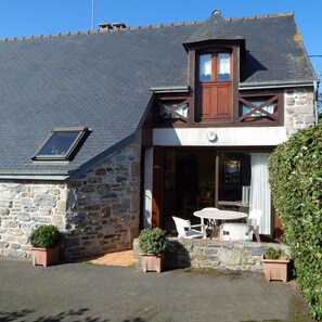 A stone building with a sloped roof, a wooden front door, and a small balcony. There are potted plants and outdoor furniture visible. - Gîtes Domaine du Grenier (Yffiniac)