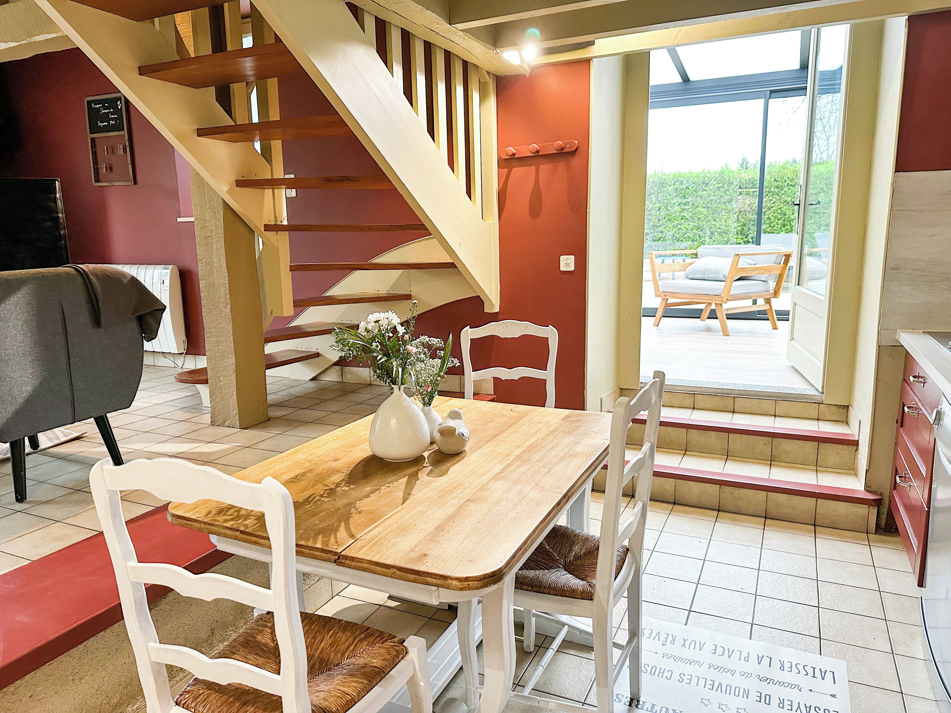 Un comedor con mesa de madera, sillas blancas, un florero con flores y una escalera que lleva a una zona de estar exterior.