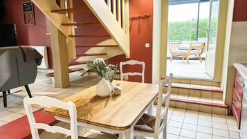Un comedor con mesa de madera, sillas blancas, un florero con flores y una escalera que lleva a una zona de estar exterior.