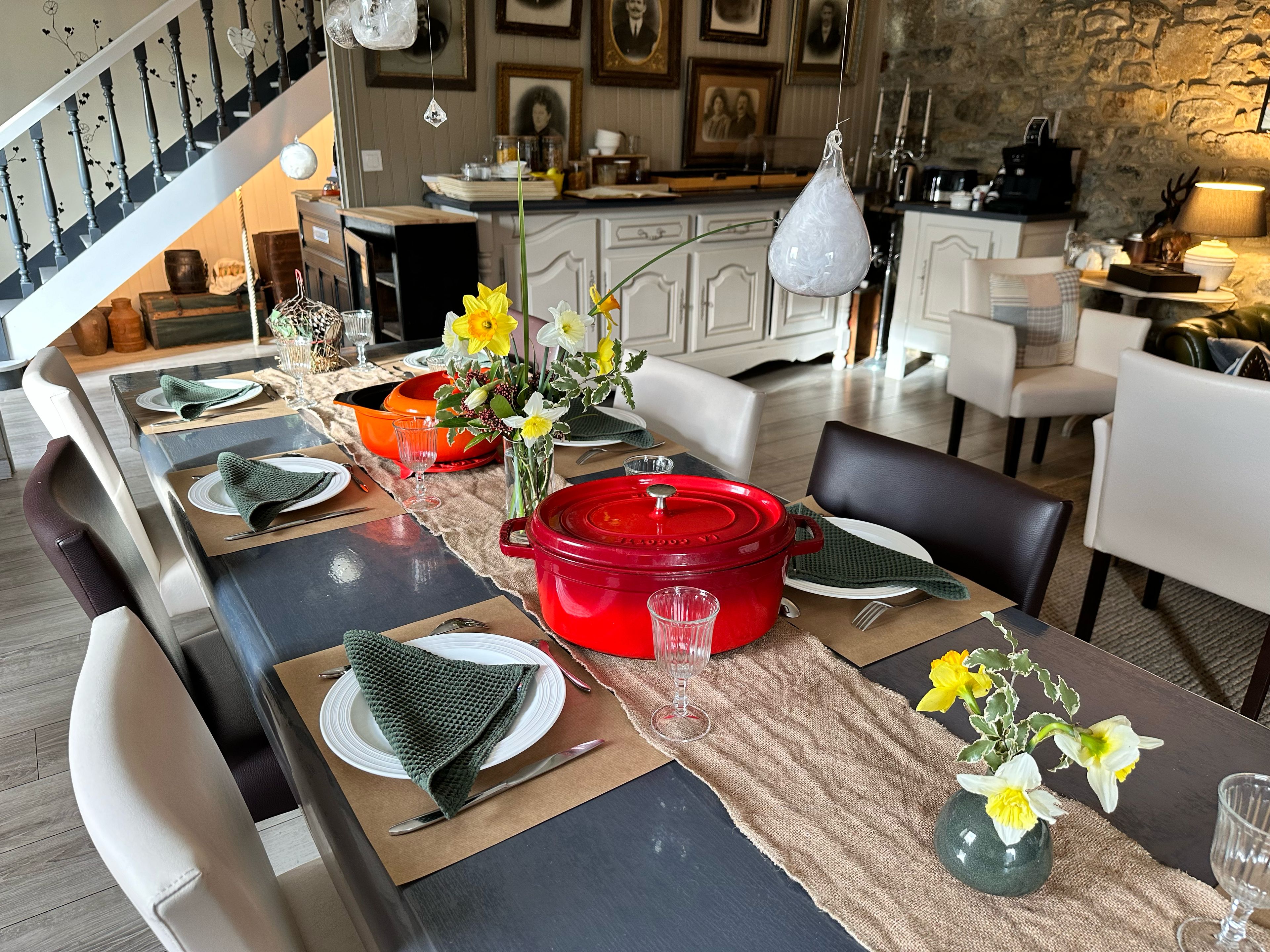 A dining area with a long table set for a meal, featuring a red pot, glassware, and flower arrangements.