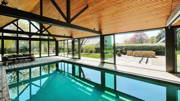 A modern indoor pool area with a wooden ceiling, large windows, and a view of the garden. - Gîtes Domaine du Grenier (Yffiniac)