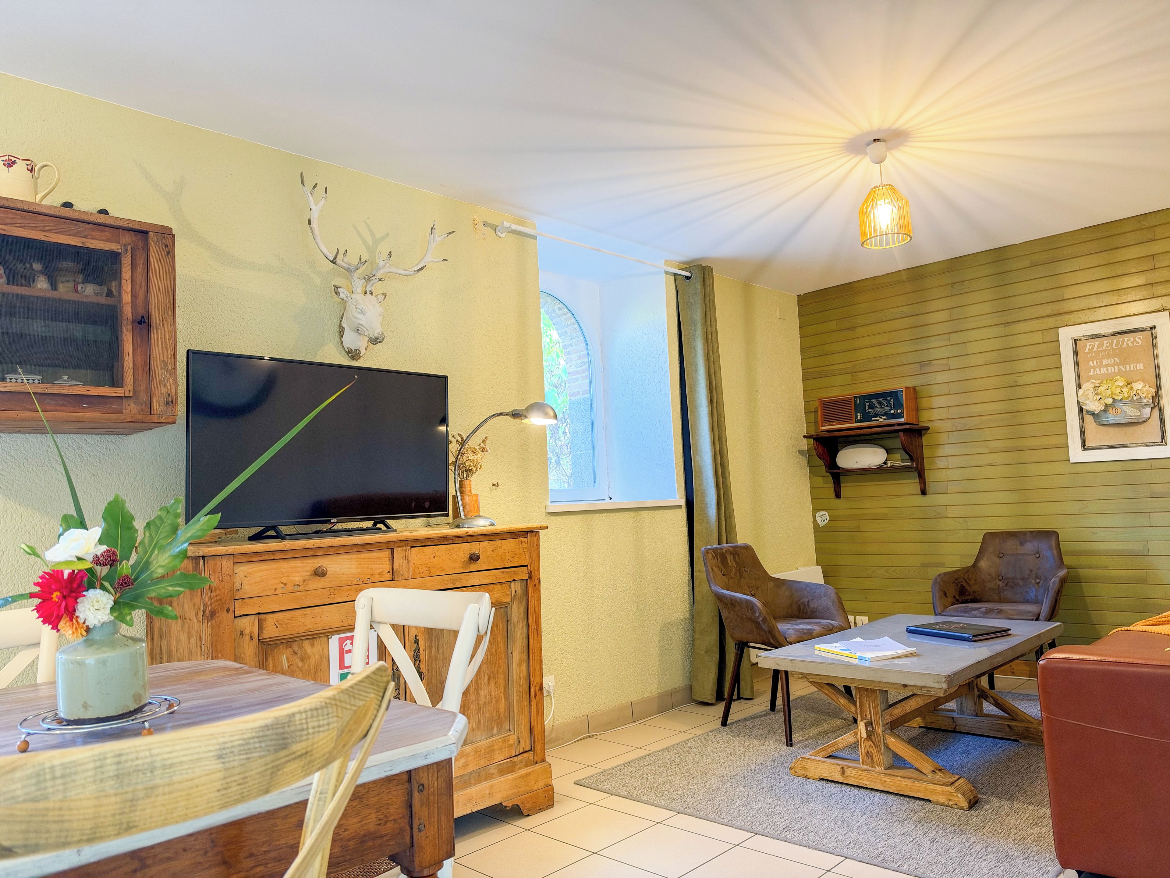 Een gezellige woonkamer met een televisie, houten meubels, een eettafel met een bloevas en een boekenplank.