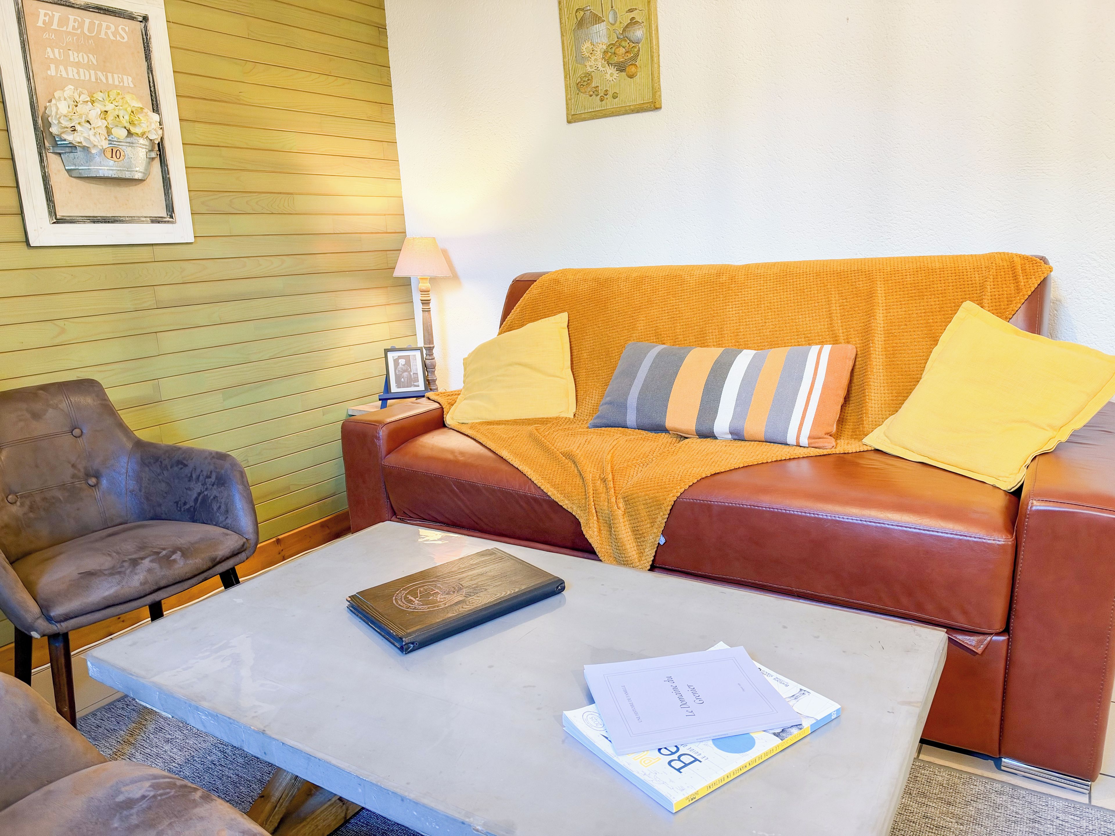 A living room with a brown leather sofa, a wooden chair, a small table with a book and papers, and a lamp.