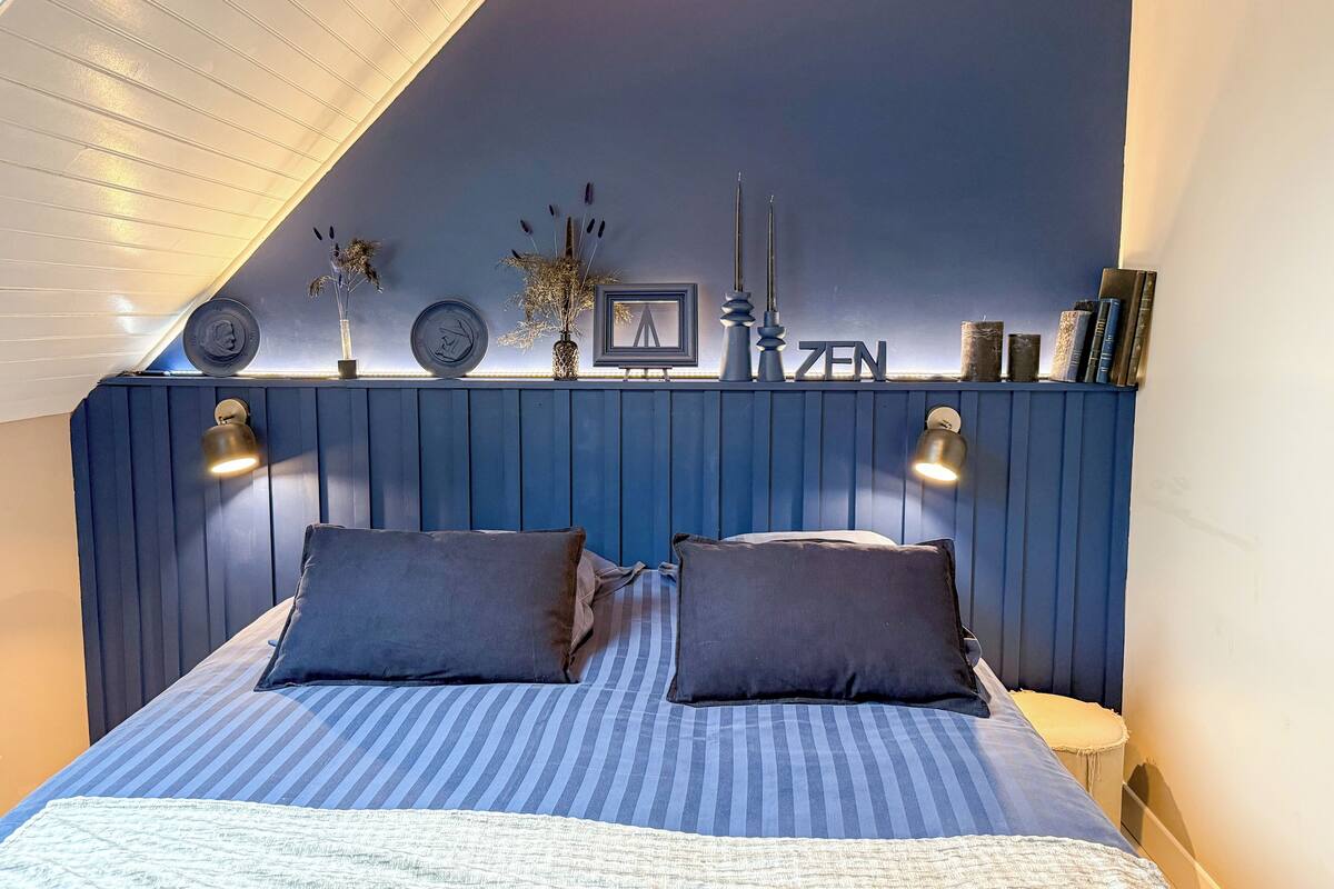 Ein Schlafzimmer mit schräg verlaufender Decke, einem Bett mit gestreifter Bettwäsche, zwei Wandlampen und einem Regal mit Dekoration.