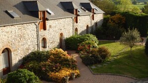 A stone building with multiple dormer windows, a paved courtyard, and surrounding greenery. - Gîtes Domaine du Grenier (Yffiniac)