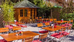 Eine Terrasse im Freien mit Holztischen und -stühlen, einer kleinen Holzkonstruktion und umgebender Bepflanzung.