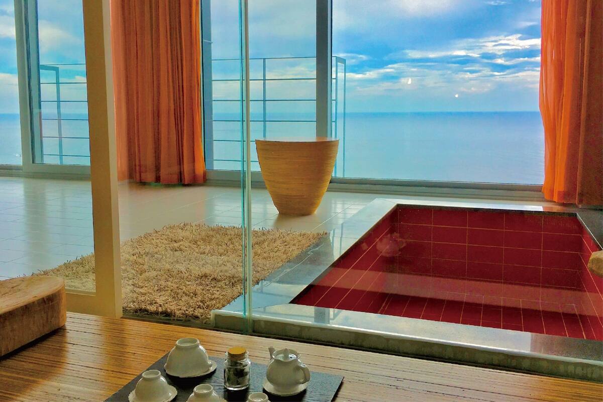 Une chambre moderne avec une grande fenêtre en verre, un bol en bois, une baignoire rouge et une table avec des théières.