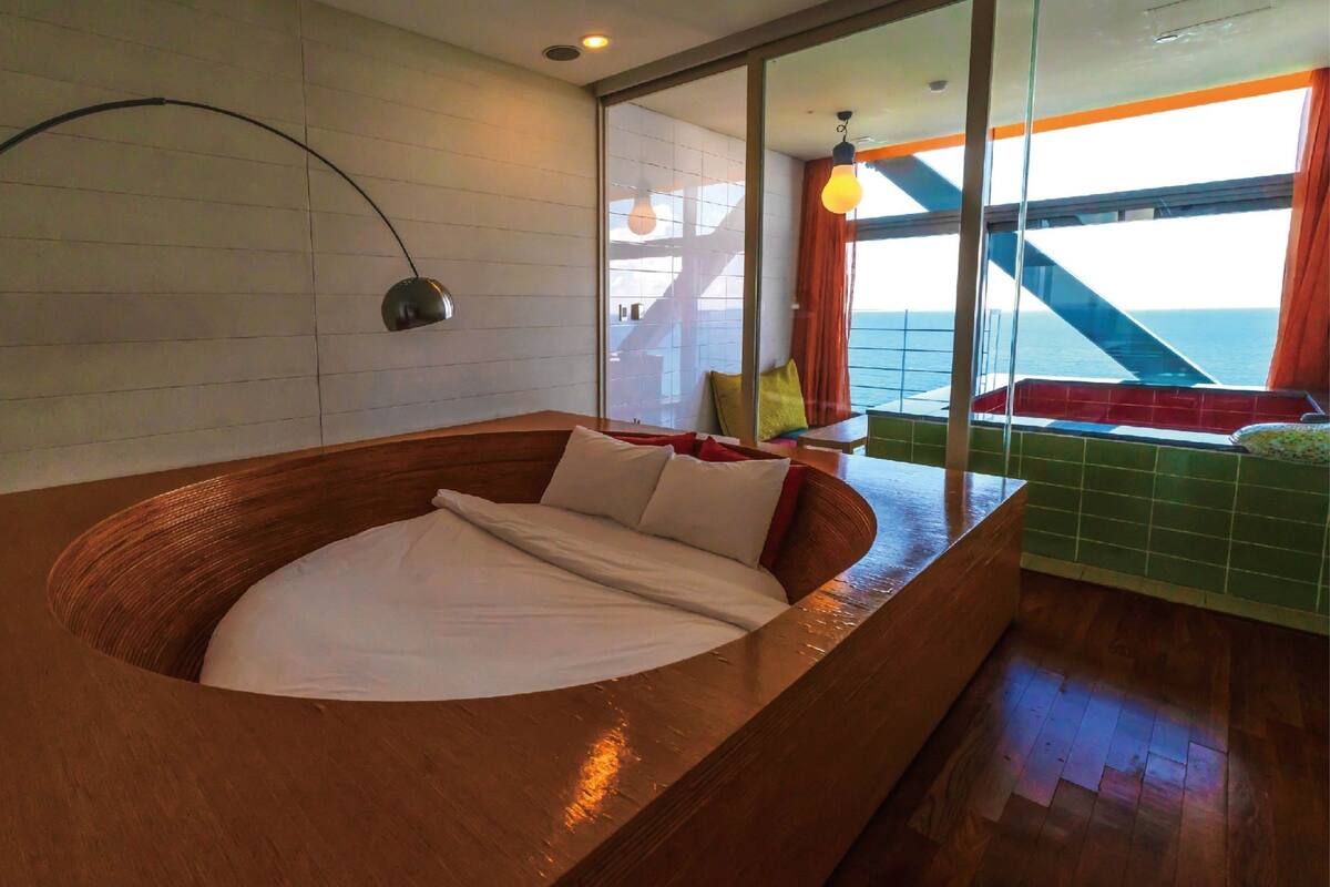 Une chambre moderne dotée d’une grande baignoire en bois, d’un banc incurvé et offrant une vue sur la mer grâce à de grandes fenêtres.