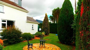 A house with a garden featuring a circular stone patio with two chairs, surrounded by various shrubs and a tall coniferous tree. - Fortview House Bed & Breakfast (Belturbet)