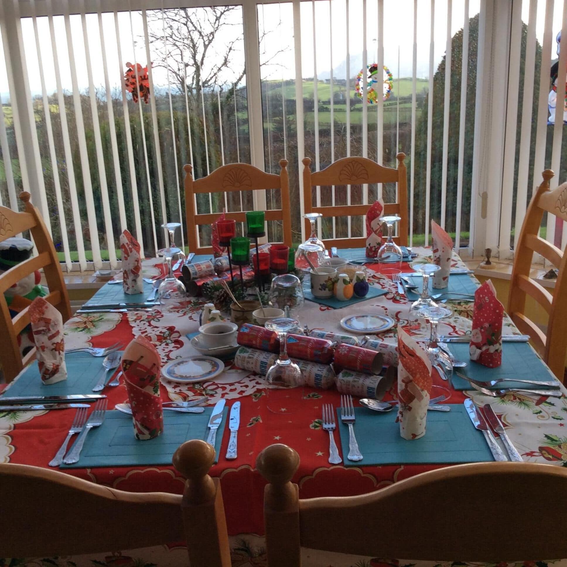 A dining table set for four with a floral tablecloth, wooden chairs, and a view of a rural landscape through large windows.