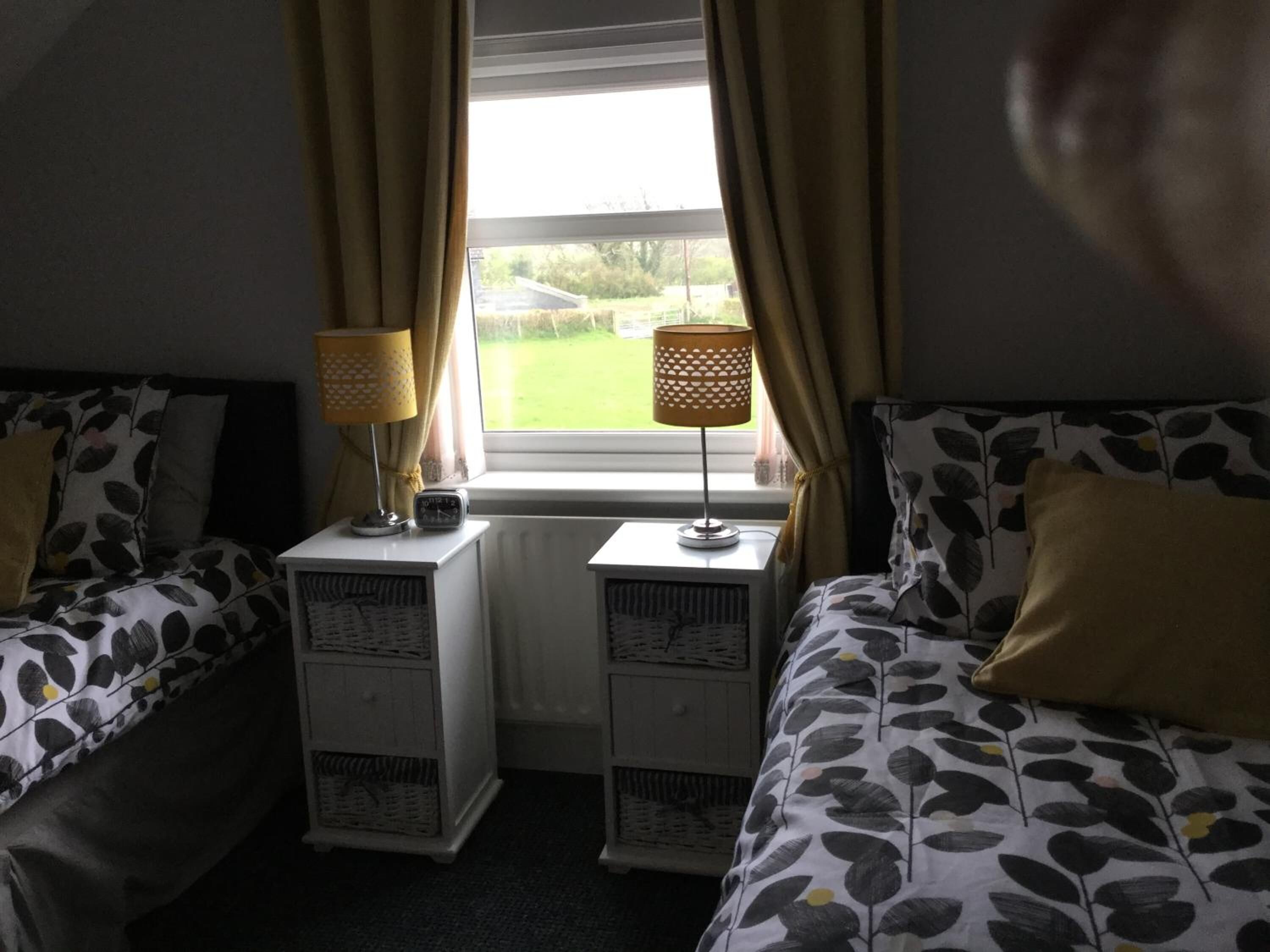A bedroom with two single beds, each with a headboard, a bedside table with a lamp, and a window with curtains.