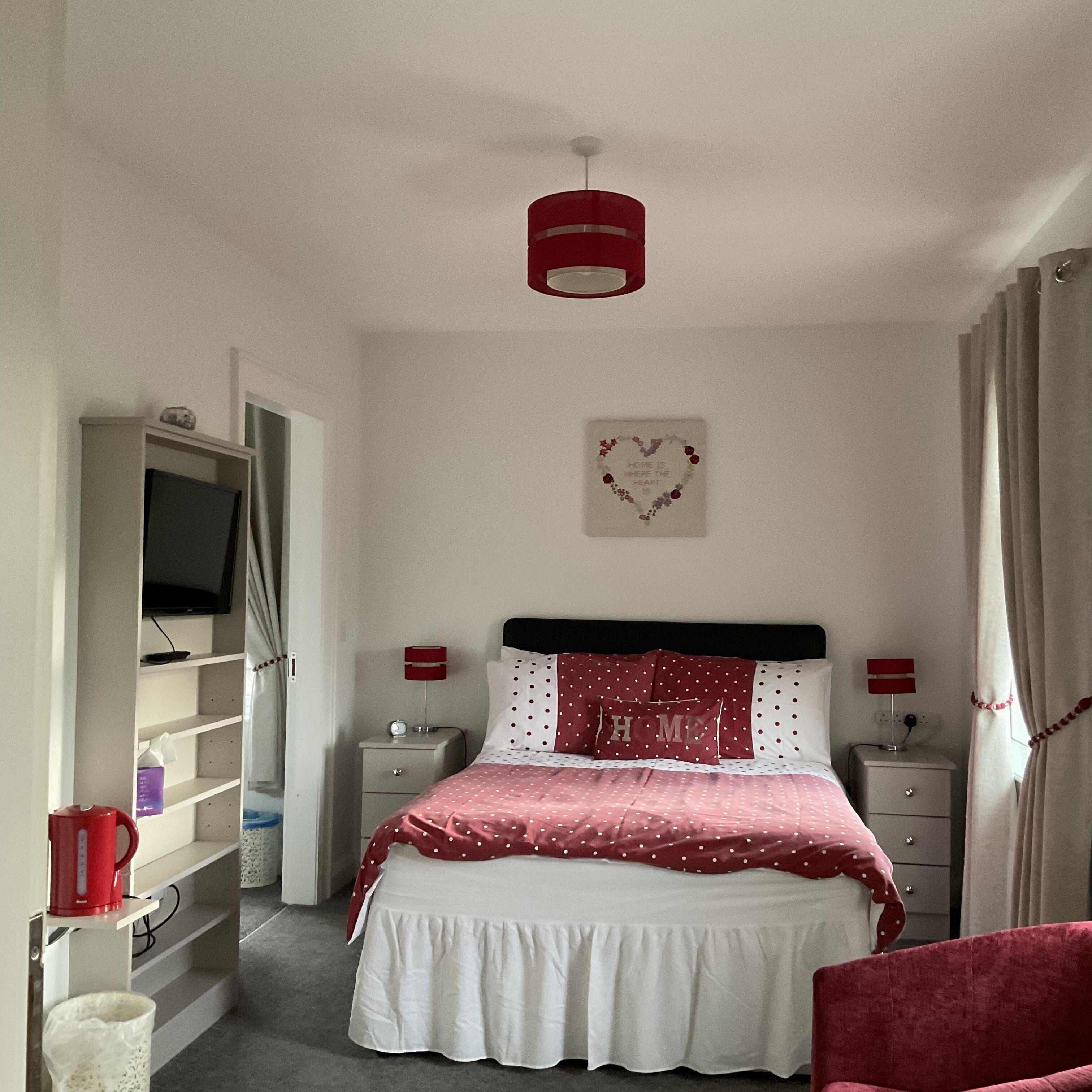 A bedroom with a bed, two bedside tables, a TV on a shelf, a red lamp, and a heart-shaped decoration on the wall.