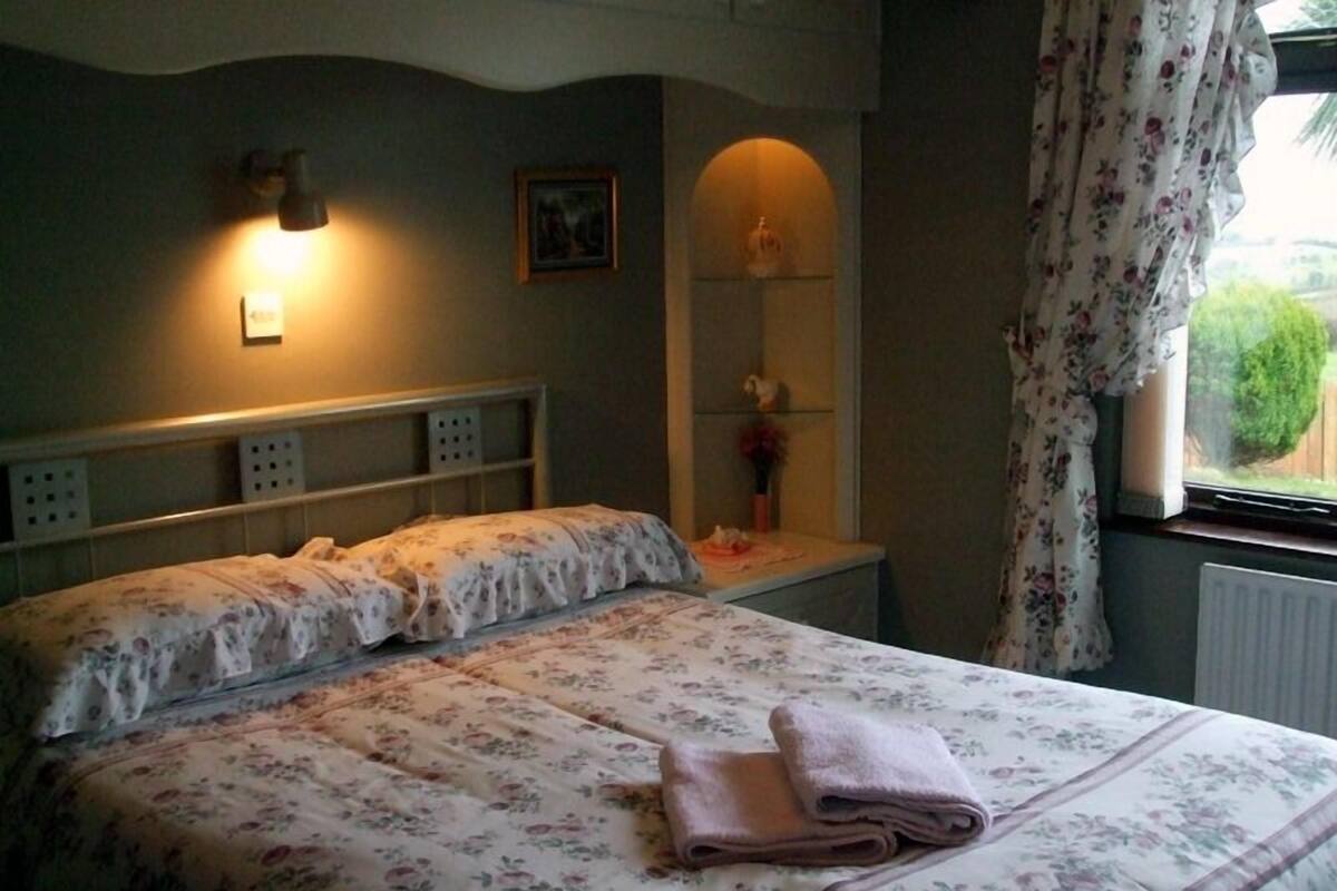 Une chambre à coucher comprenant un lit, une table de chevet, une lampe fixée au mur, un tableau, une étagère avec des objets décoratifs et une fenêtre avec des rideaux.