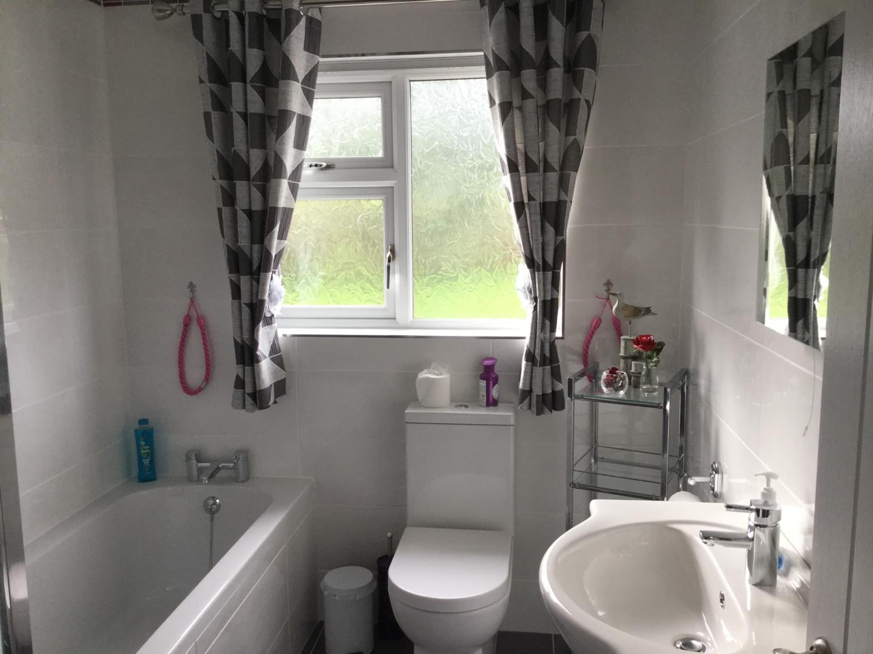 A bathroom with a bathtub, toilet, and sink. There is a window with curtains, a towel rack, and a small shelf with toiletries.