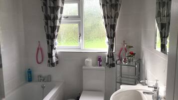 A bathroom with a bathtub, toilet, and sink. There is a window with curtains, a towel rack, and a small shelf with toiletries.