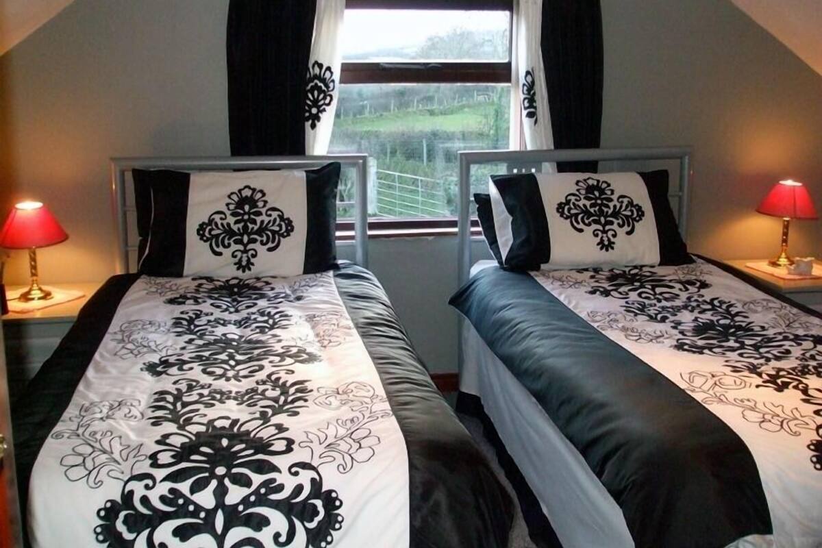 Une chambre double avec deux lits simples, chacun recouvert d’une couvre-lit à motifs noir et blanc et agrémenté de coussins assortis. On y trouve deux tables de chevet avec des lampes, ainsi qu’une fenêtre avec des rideaux noirs.