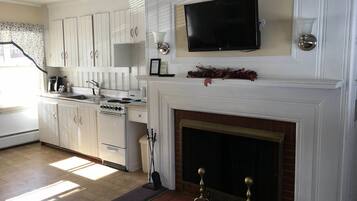 A kitchen with white cabinets, a stove, a sink, a television above the fireplace, and a mantel with decorations.