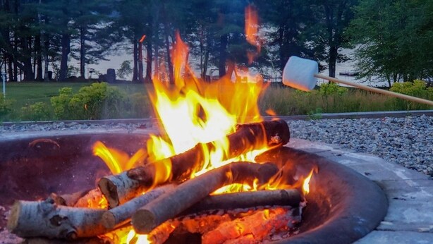A campfire with logs and a shovel in a metal fire pit surrounded by gravel. - Highland Lake Resort (Bridgton)