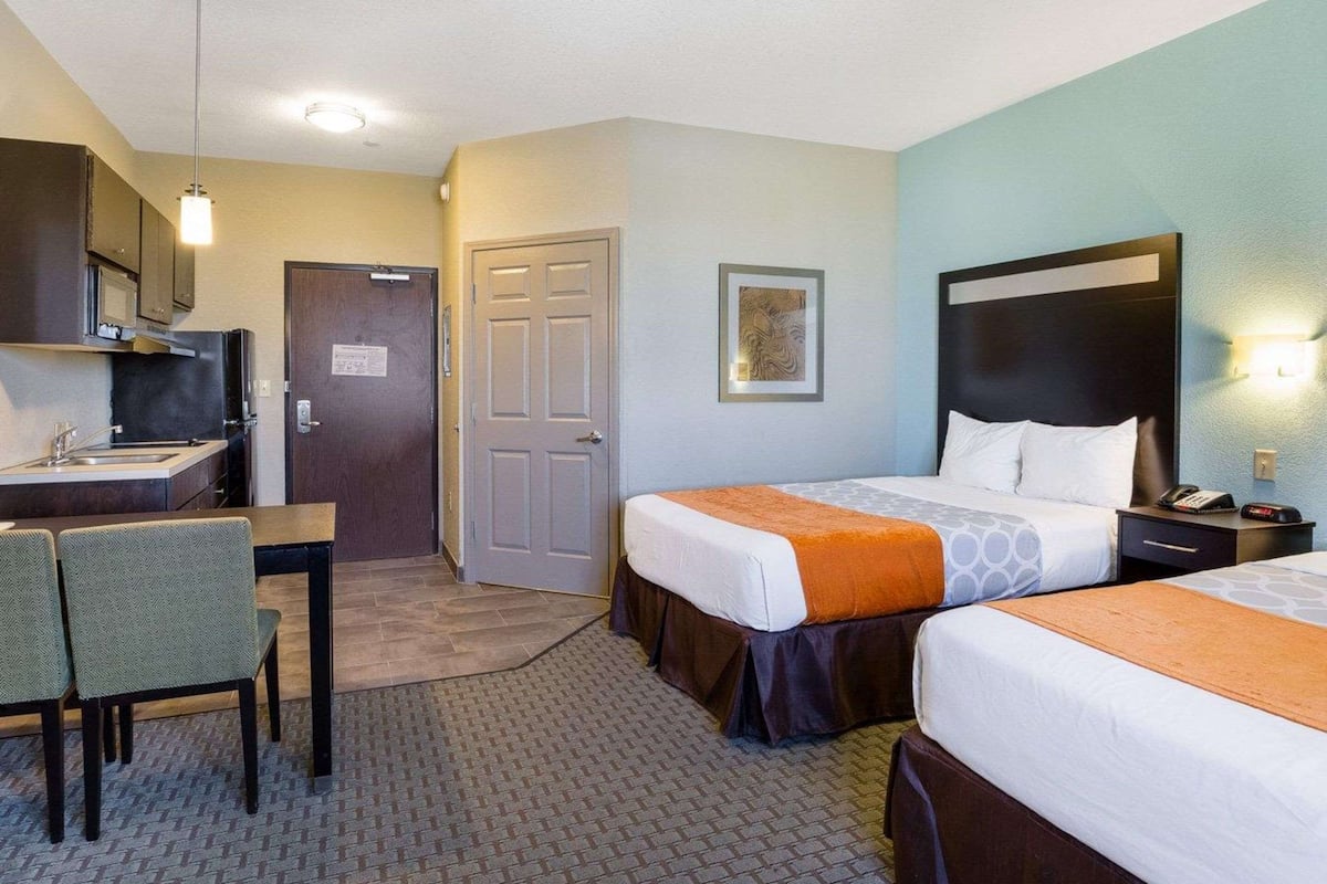A hotel room with two beds, a desk with chairs, a kitchenette, and a door.