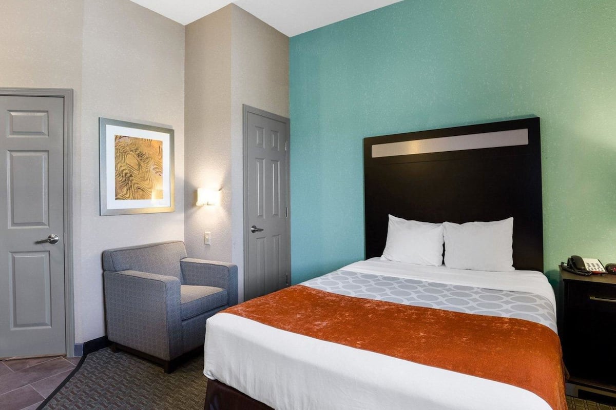 A hotel room with a bed, a chair, a desk with a phone, and a door.