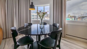 A modern dining area with a round table, six chairs, a vase with flowers, and a view of buildings through large windows. - Hlemmur Square (Reykjavik)