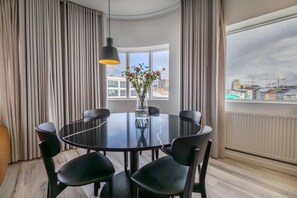 A modern dining area with a round table, six chairs, a vase with flowers, and a view of buildings through large windows. - Hlemmur Square (Reykjavik)