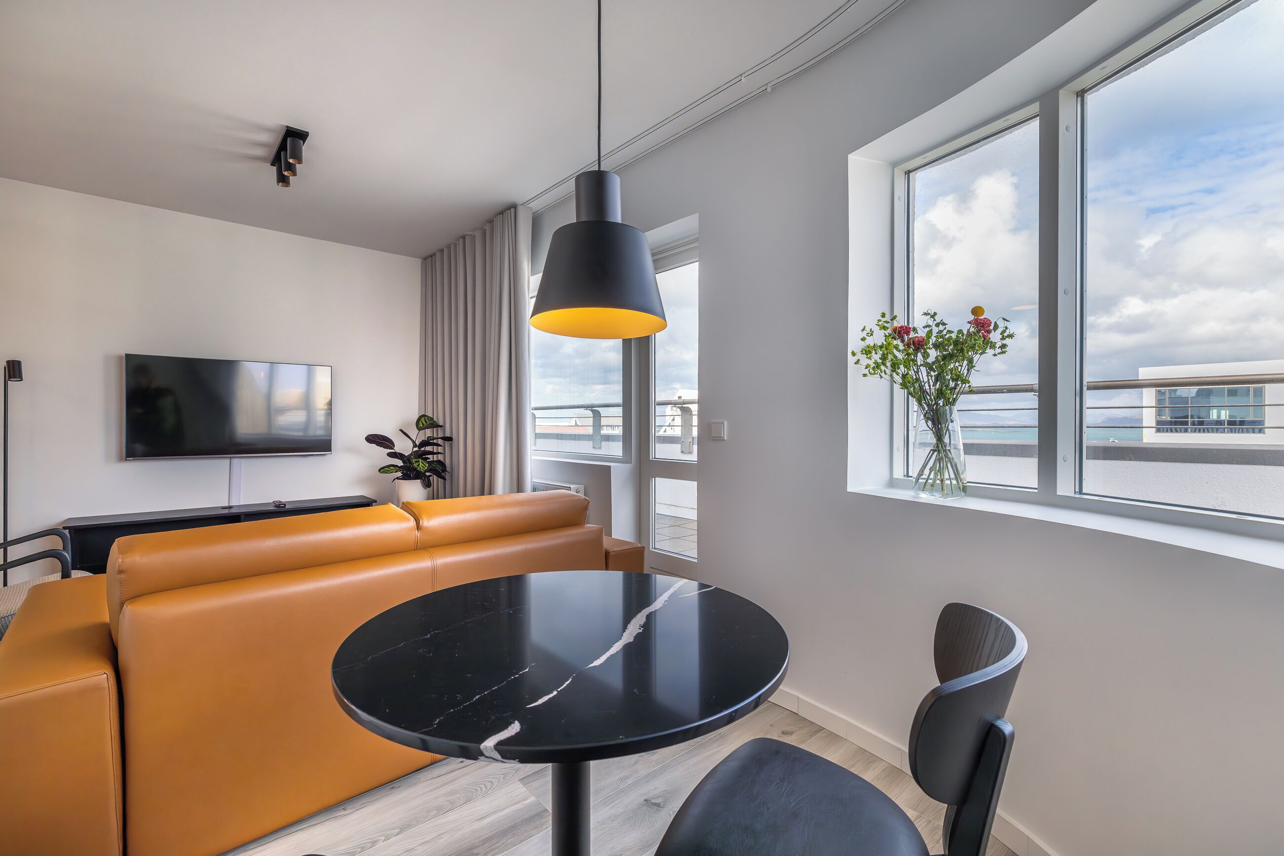 Una sala de estar moderna con una mesa de mármol negro, un sofá de cuero marrón, una lámpara colgante negra, un televisor de pantalla plana y un ventanal con vistas a la ciudad.