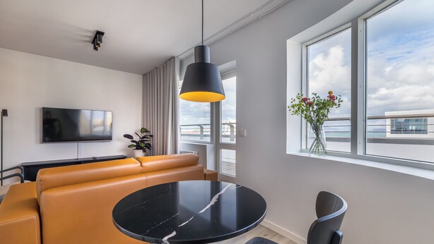 A modern living room with a black marble table, a brown leather sofa, a black pendant light, a flat-screen TV, and a large window with a view of the city. - Hlemmur Square (Reykjavik)