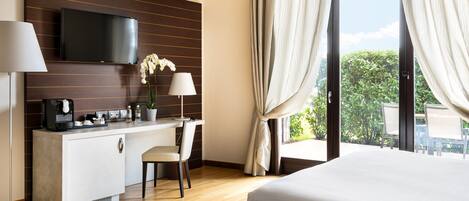 Ein Hotelzimmer mit Bett, Schreibtisch mit Flachbildfernseher, Kaffeemaschine, Zimmerpflanze und Blick ins Grüne durch das Fenster.