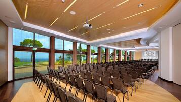 Un salón de conferencias con hileras de sillas, amplias ventanas con vistas y un techo de madera con iluminación empotrada.