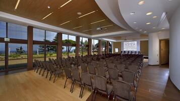 Uma sala de conferências com fileiras de cadeiras, grandes janelas e uma tela para apresentações.