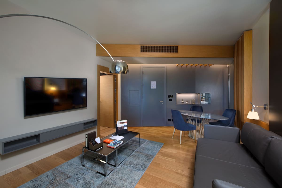 Ein modernes Wohnzimmer mit Flachbildfernseher, Couchtisch und Sofa. Es gibt eine Essecke mit Tisch und Stühlen, und im Hintergrund eine Küche.