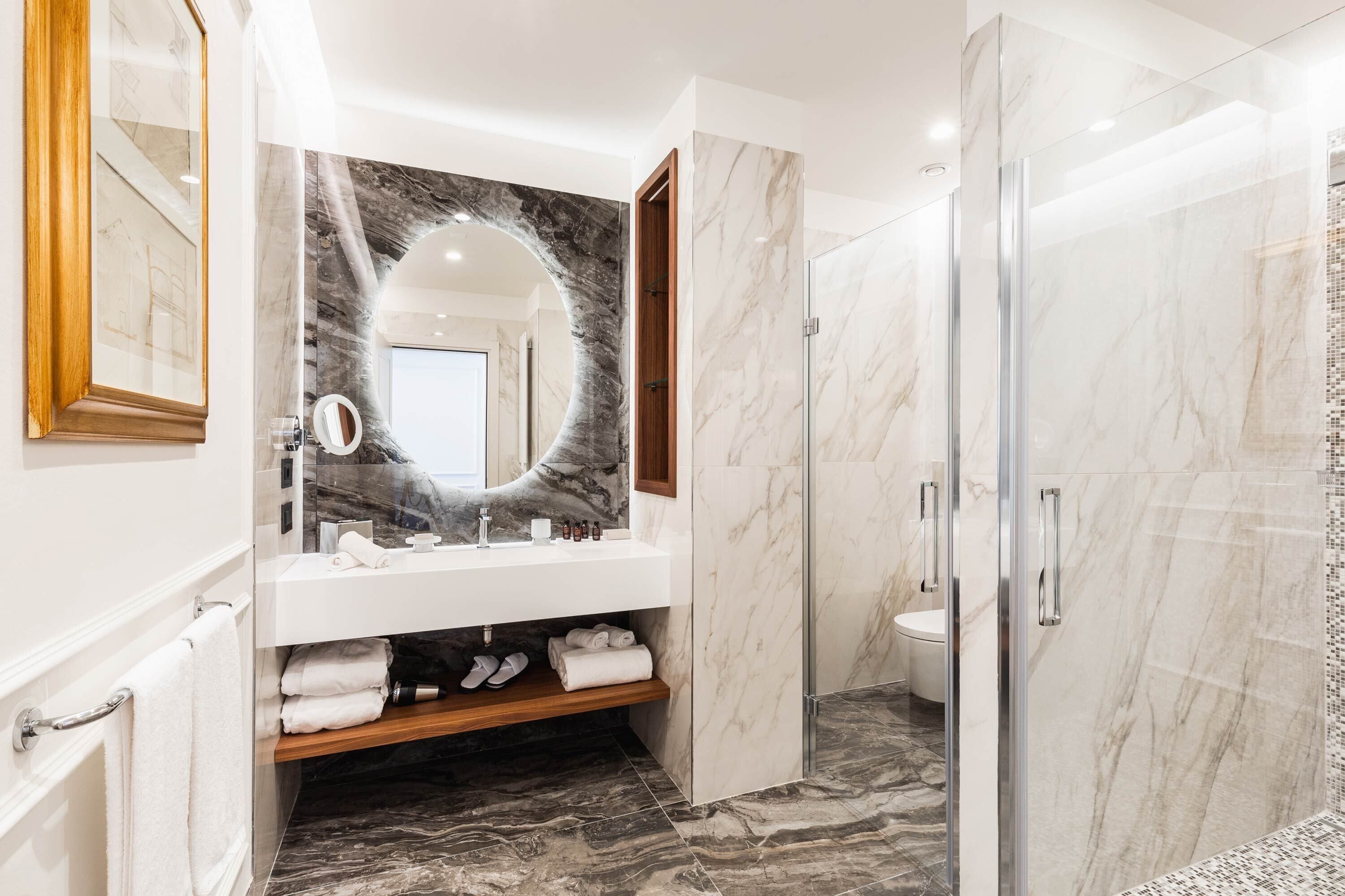 A modern bathroom with a marble vanity, a glass-enclosed shower, and a wooden shelf with towels.