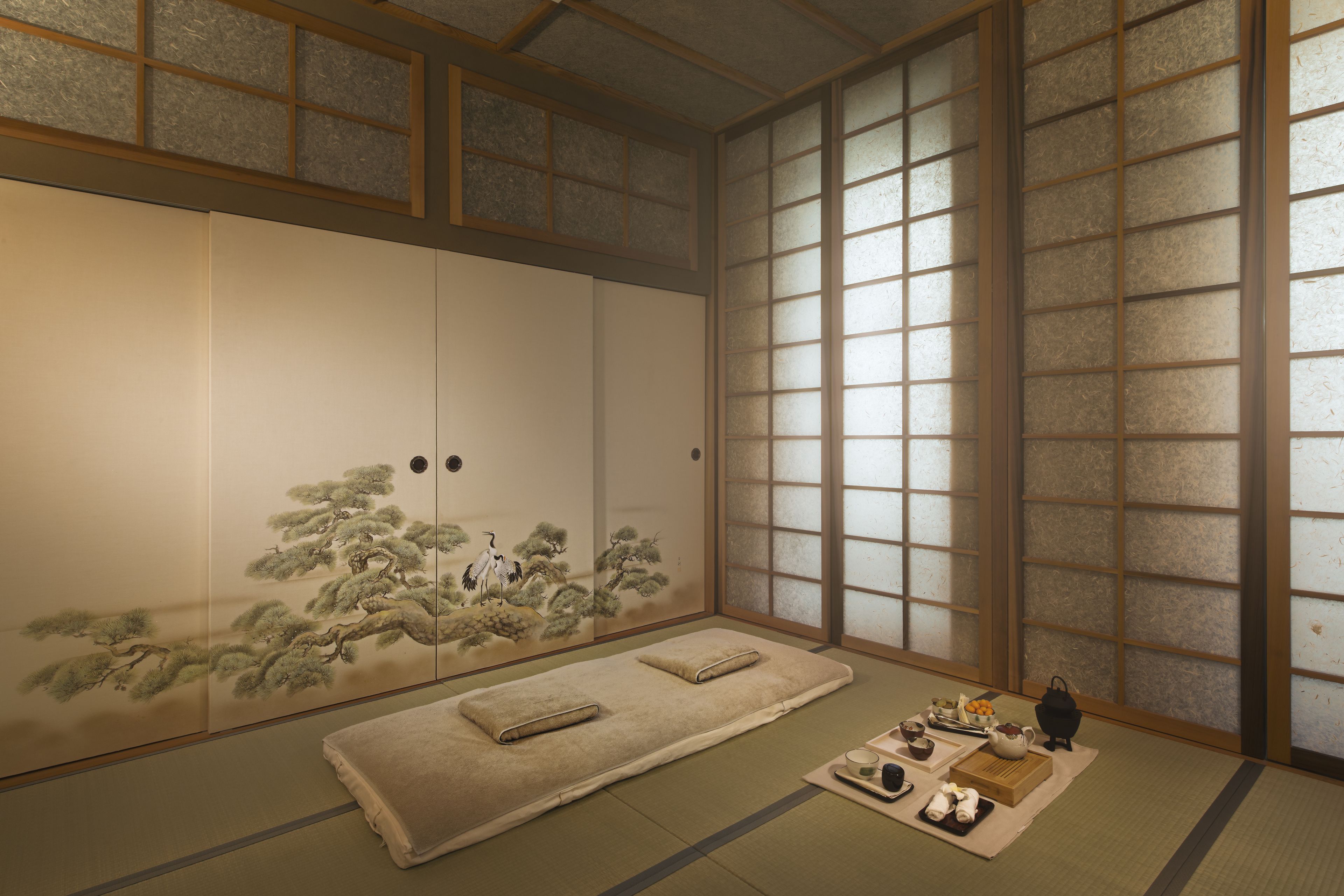 Una habitación japonesa tradicional con suelo de tatami, puertas correderas con una pintura de motivos naturales y un juego de té sobre una mesa baja.