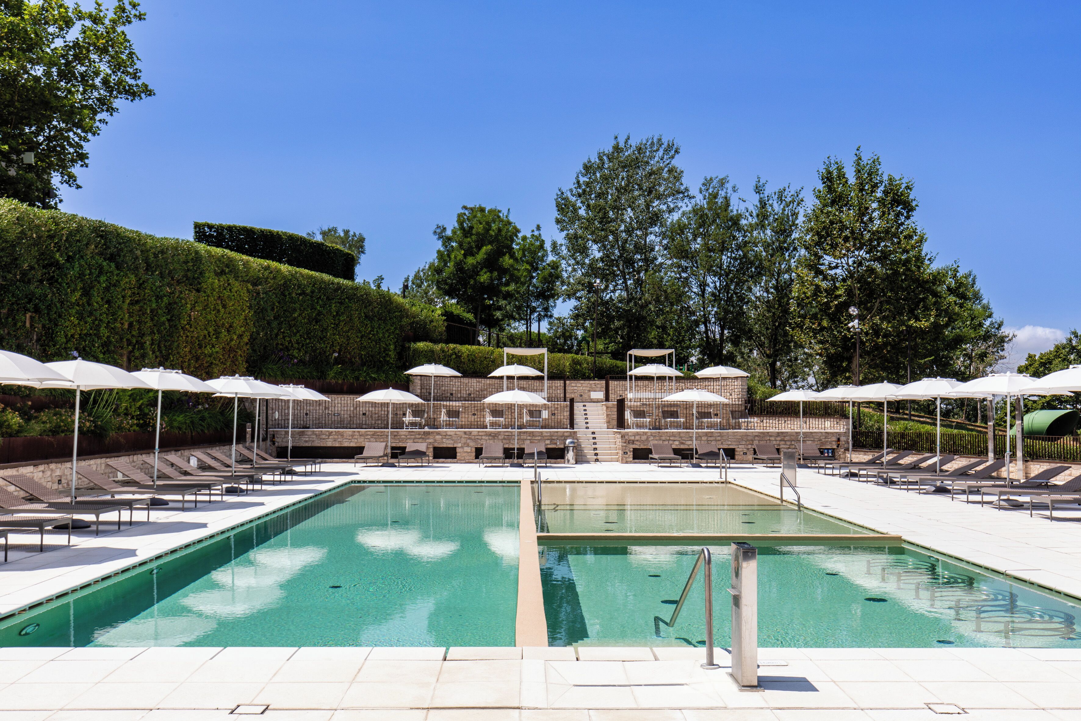 A swimming pool with lounge chairs and umbrellas, surrounded by a landscaped garden and a building with steps leading up to it.