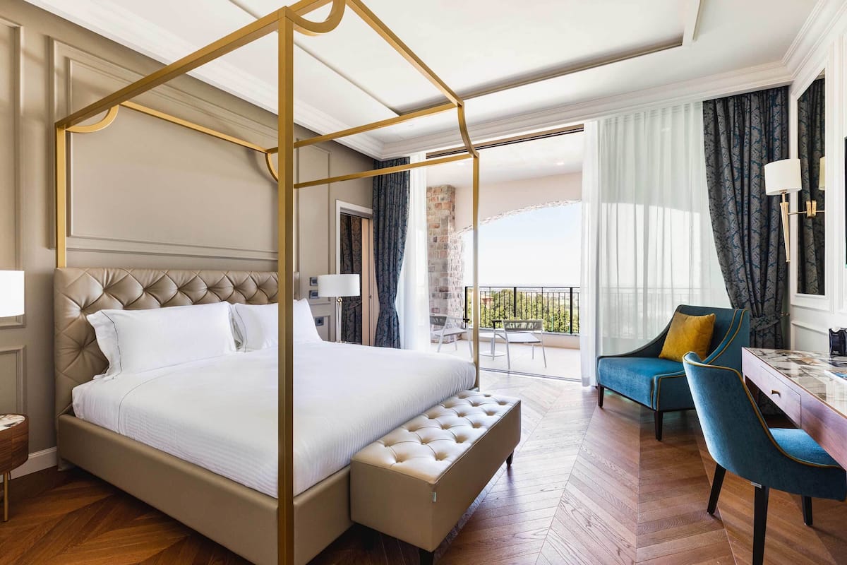 Ein geräumiges Schlafzimmer mit Himmelbett, eine Sitzecke mit blauem Sessel und ein Balkon mit Aussicht.
