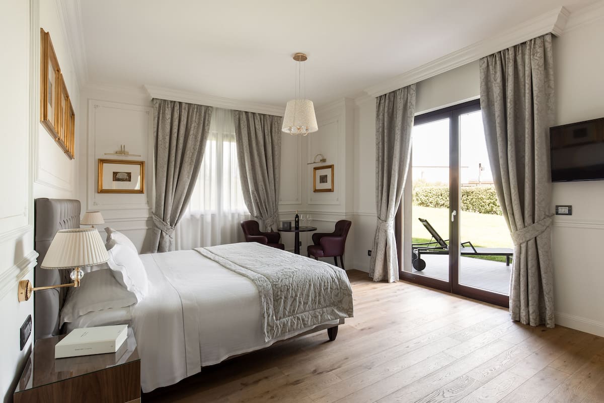 Ein Hotelzimmer mit Bett, Nachttisch mit Lampe, Wand-Fernseher, einem kleinen Tisch mit Stühlen und Blick durch die Glastür.