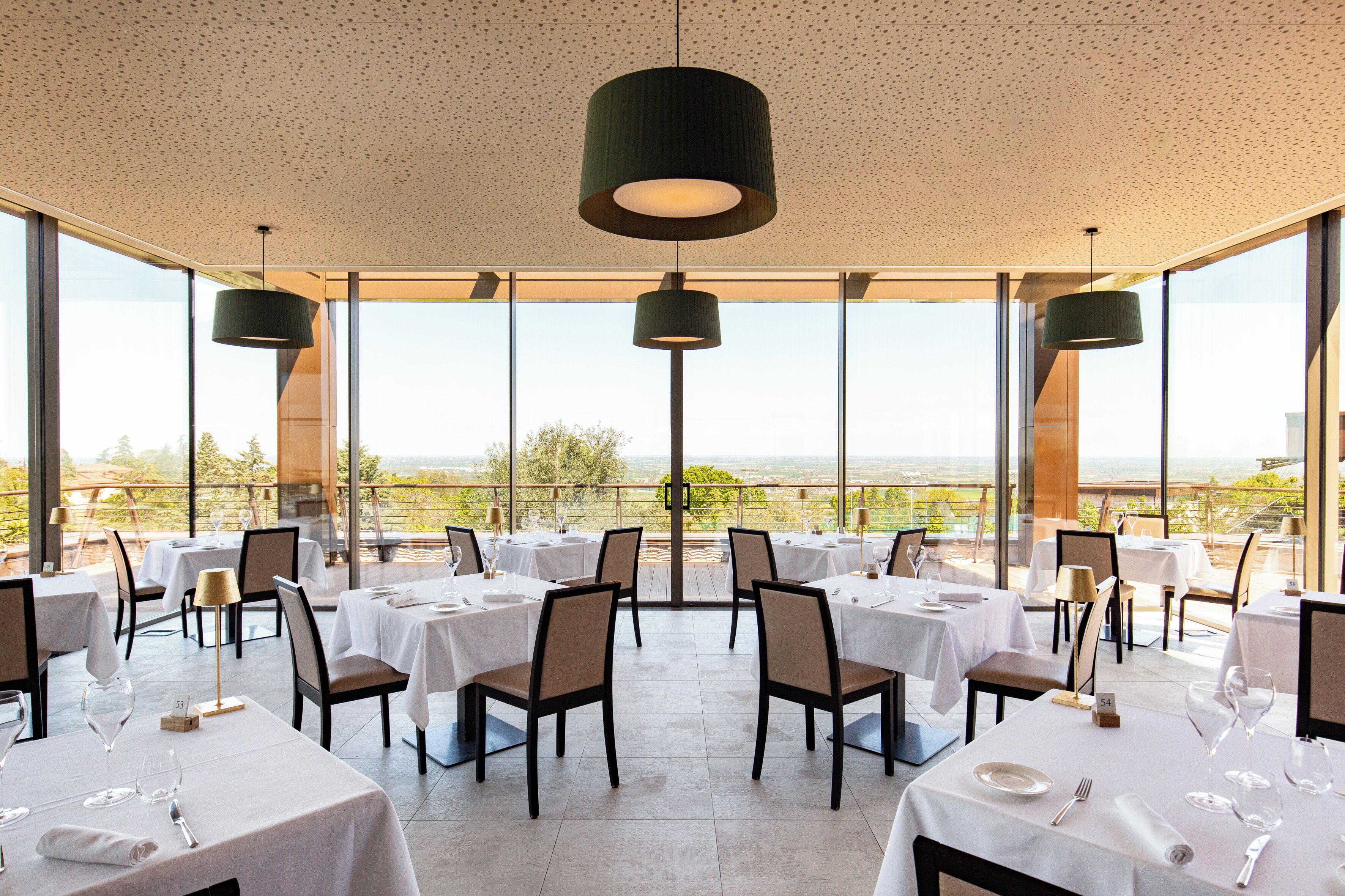 A restaurant with tables set for dining, large windows offering a view, and hanging pendant lights.