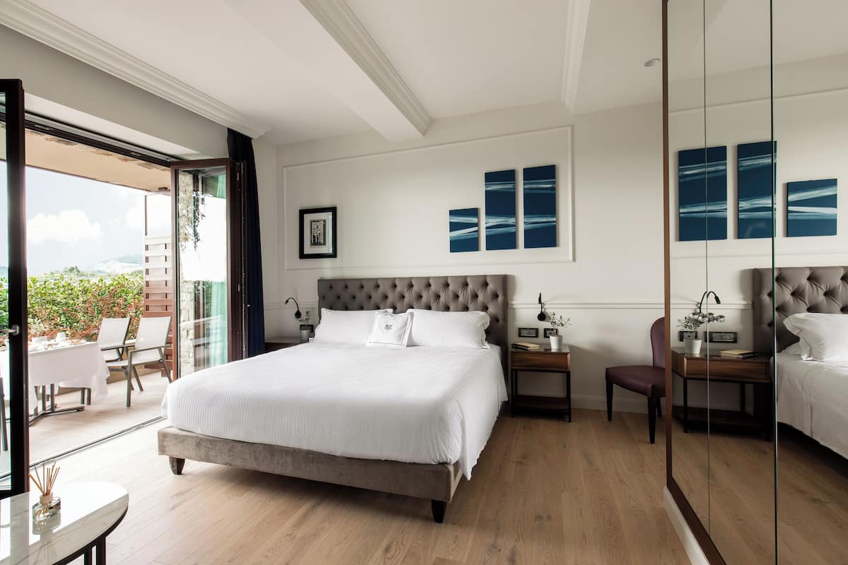 Ein Schlafzimmer mit einem großen Bett, einem Nachttisch, einem Sessel und Blick ins Freie.
