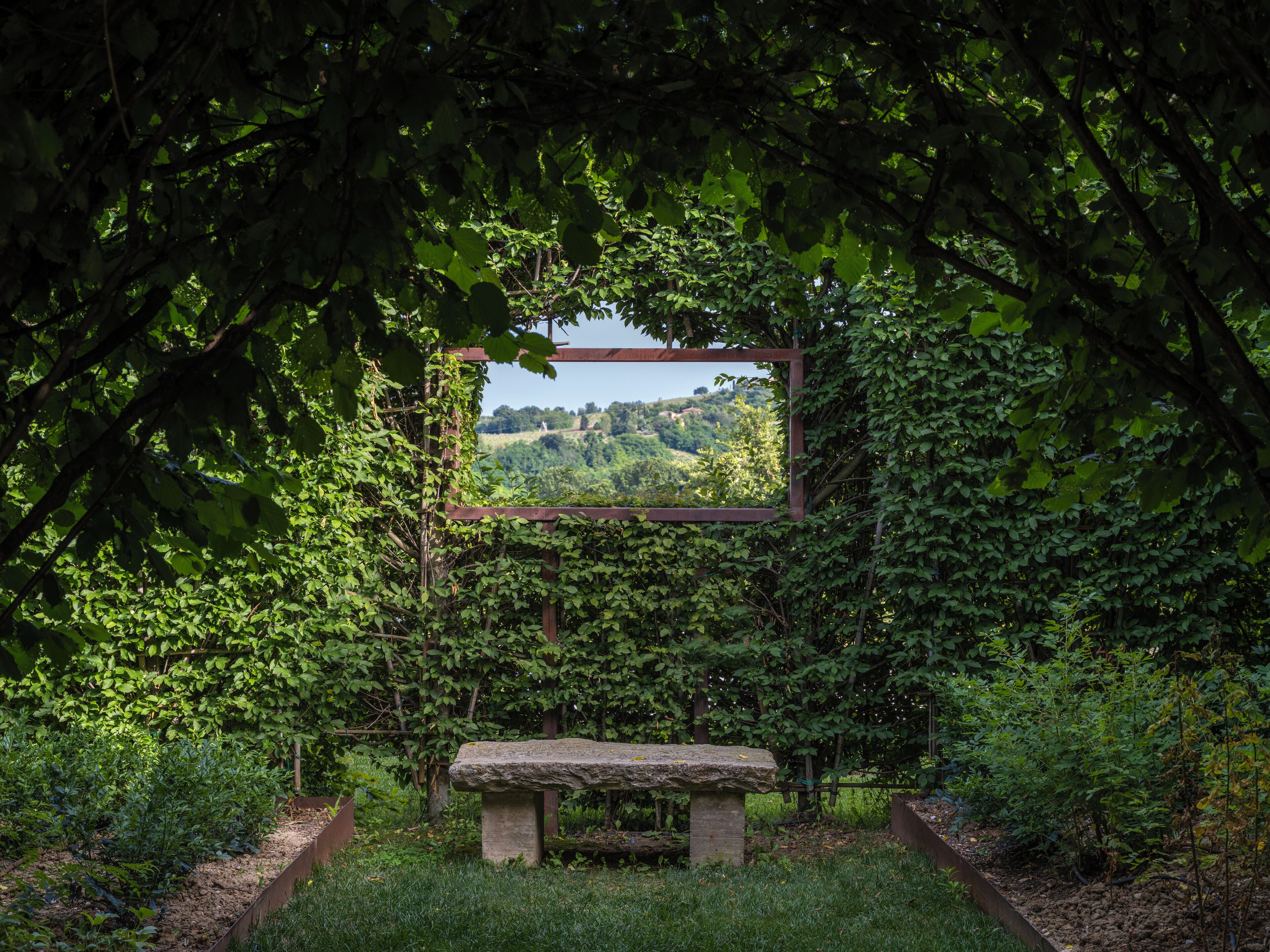 A stone bench surrounded by lush greenery and a wooden pergola framing a view of a hilly landscape.