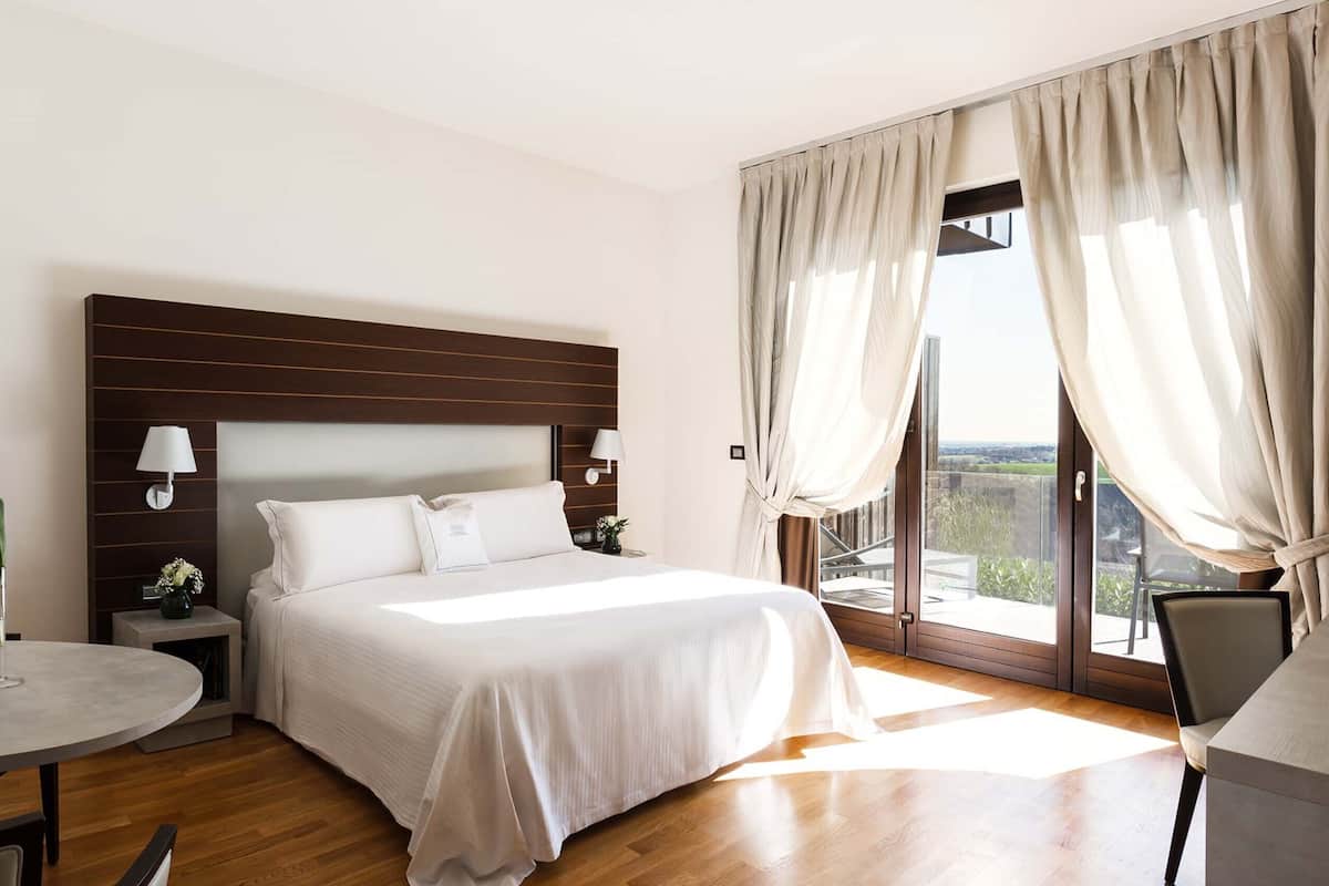 Ein Hotelzimmer mit einem großen Bett, einem Nachttisch mit Lampe, einem kleinen Tisch mit Vase und Blick ins Freie durch das Fenster.